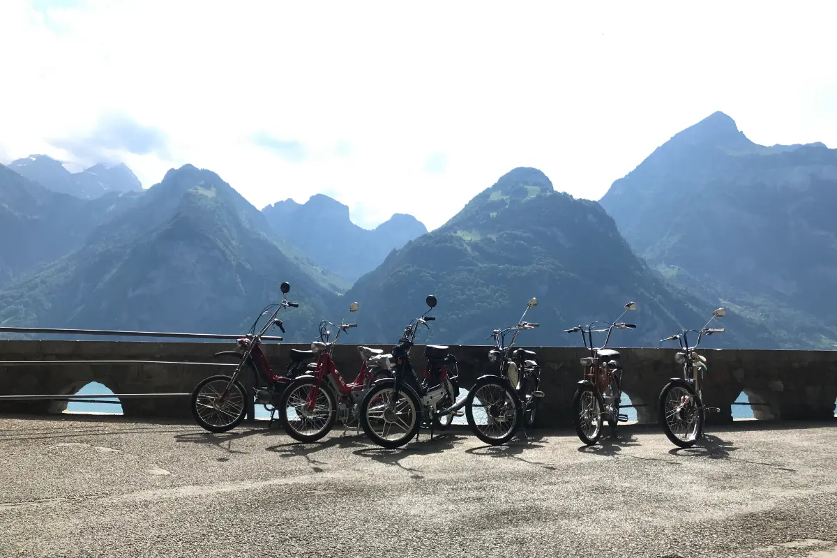 Sechs Töffli in einer Reihe an einer Mauer über dem See, dahinter steile Berge.